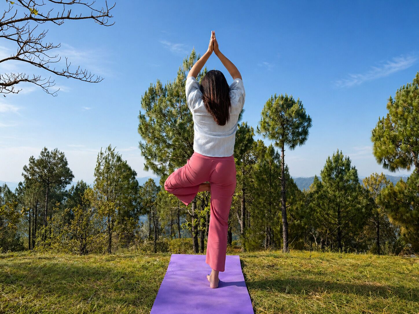 Una persona practicando yoga sobre una colchoneta morada al aire libre, con árboles y un cielo despejado de fondo.