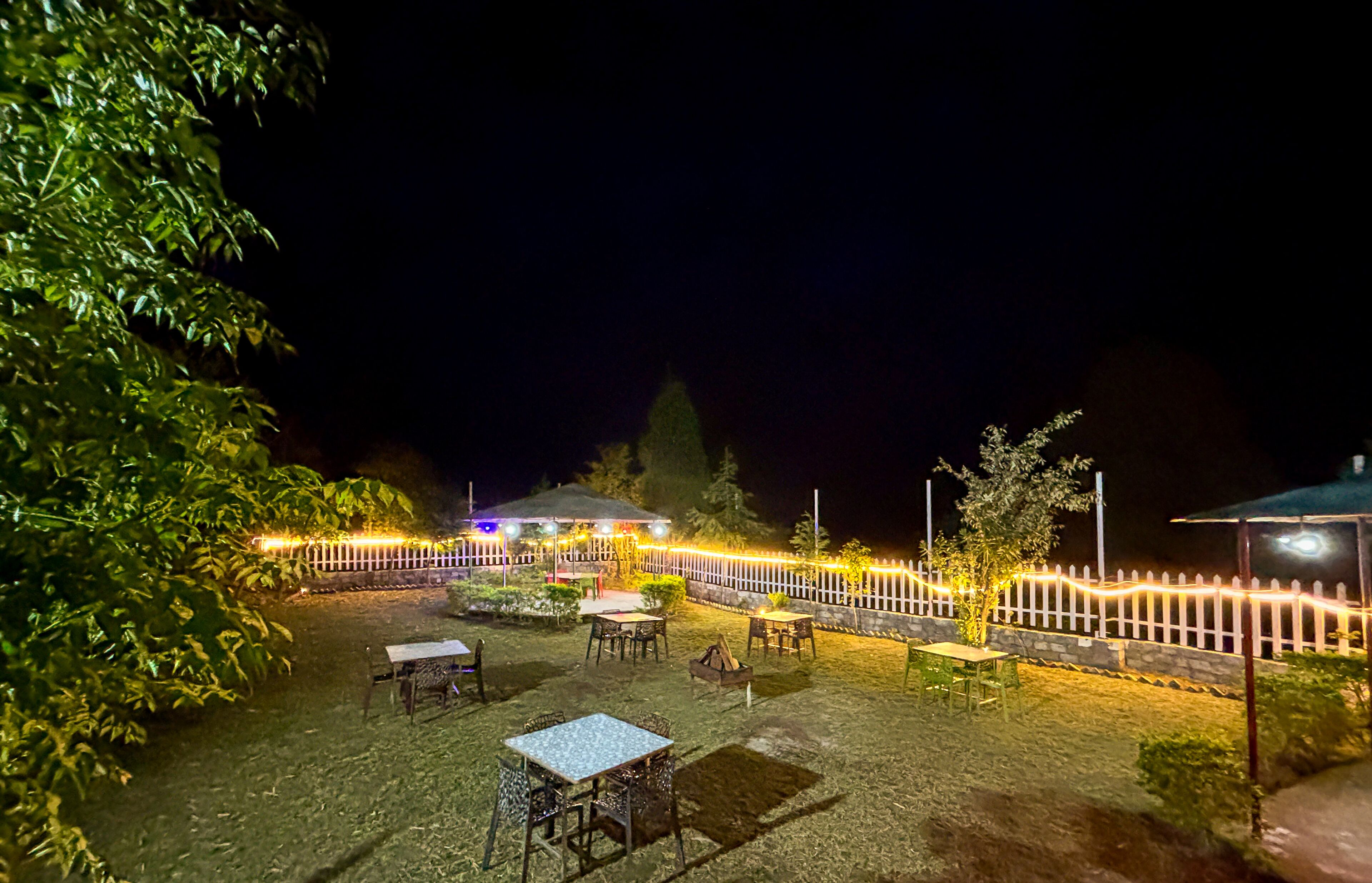 Een terras buiten met tafels en stoelen, een witte hek en sfeerverlichting.
