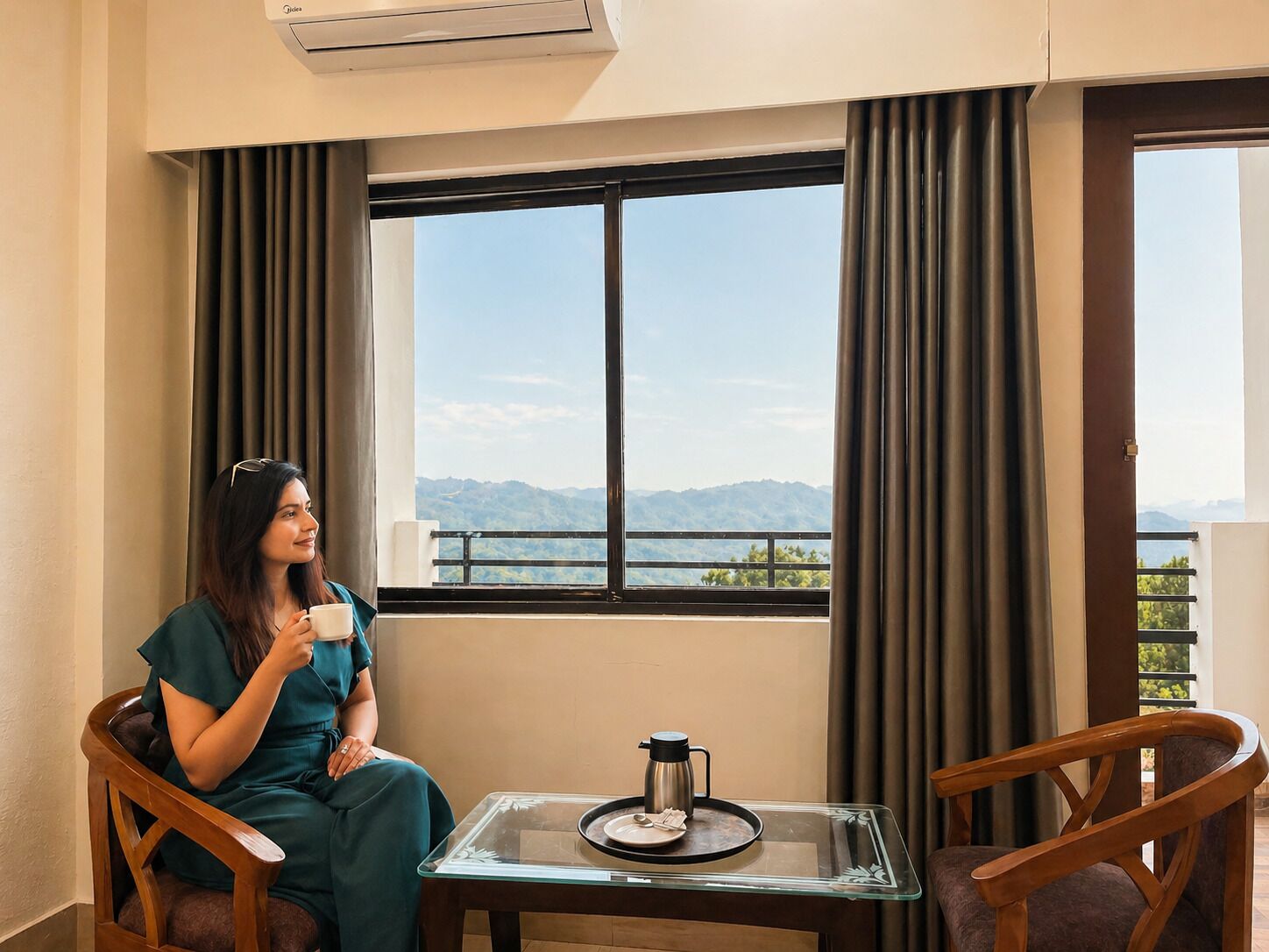 Una habitación con vistas a montañas, una mesa con una cafetera y una persona sentada junto a la ventana.