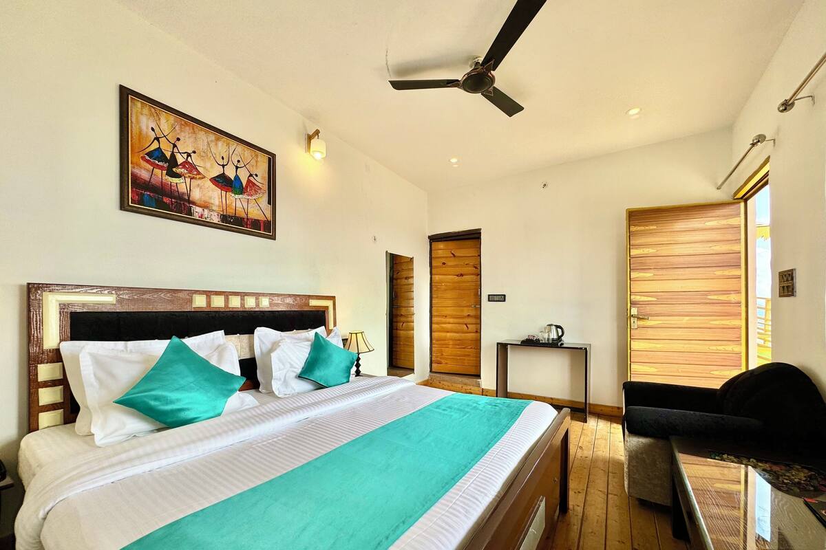A hotel room with a large bed, a ceiling fan, a wooden door, and a small table with a kettle.