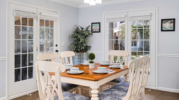 Un comedor con una mesa de madera dispuesta para cuatro personas, rodeada de sillas blancas con cojines azules. Hay dos ventanas grandes con marcos blancos, una planta en maceta y un pequeño objeto decorativo sobre la mesa.