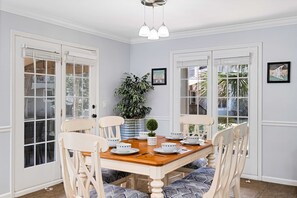 Un comedor con una mesa de madera dispuesta para cuatro personas, rodeada de sillas blancas con cojines azules. Hay dos ventanas grandes con marcos blancos, una planta en maceta y un pequeño objeto decorativo sobre la mesa.