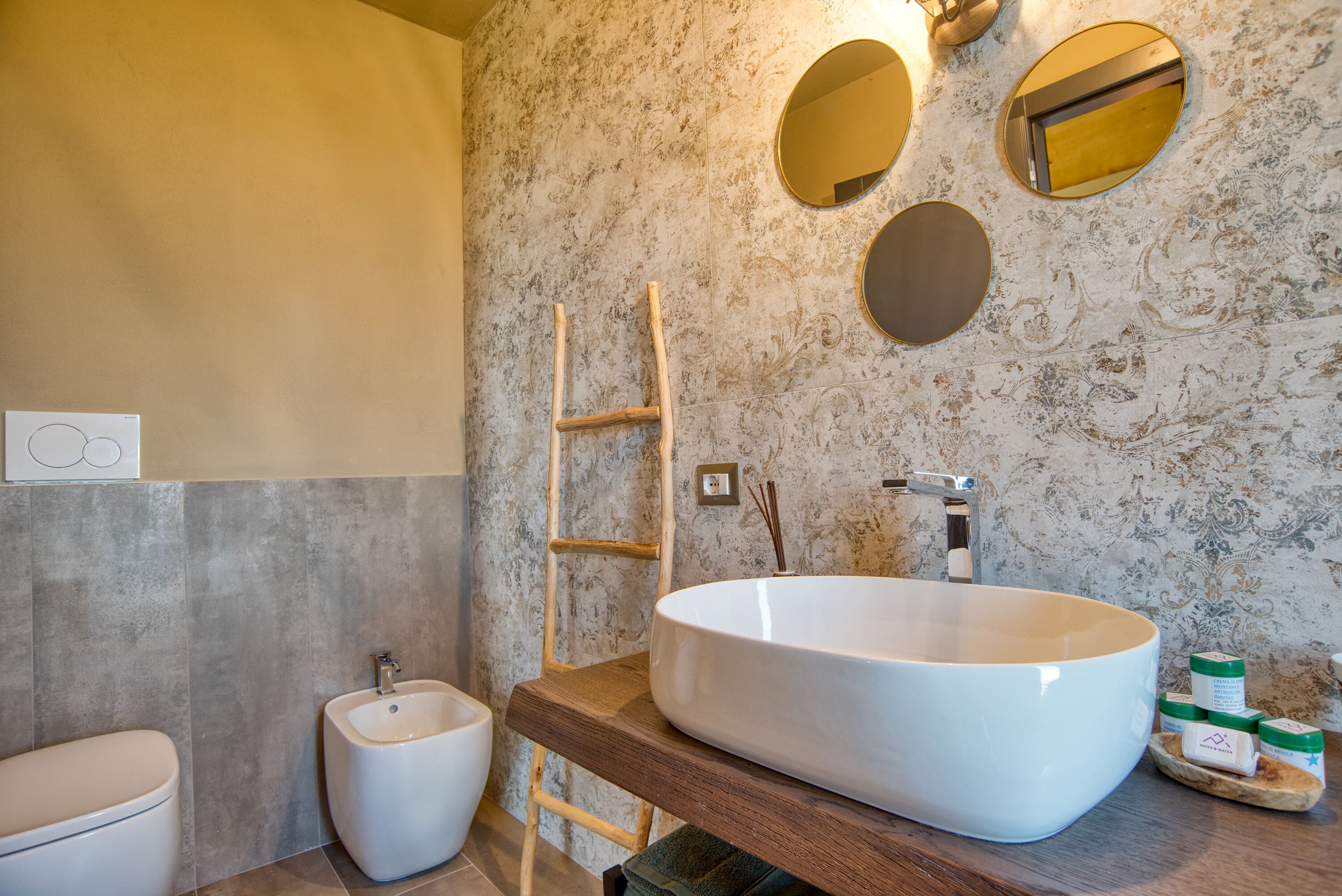 Ein Badezimmer mit einem großen weißen Waschbecken, einer Holzleiter, einem Bidet und einer Marmorwand mit dekorativen Spiegeln.