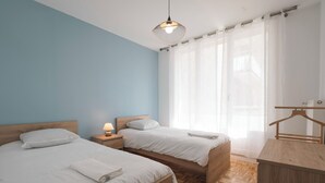 A bedroom with two beds, a nightstand with a lamp, a wooden dresser, and a window with sheer curtains. - #HV | Le Panorama 🏔️ (Saint-Martin-d'Hères)
