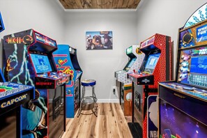 A room with arcade games, a wooden floor, and a wall-mounted poster. - Luxe 6BR Pool Cabin*Hot Tub*Arcade*Sleeps 20 (Sevierville)