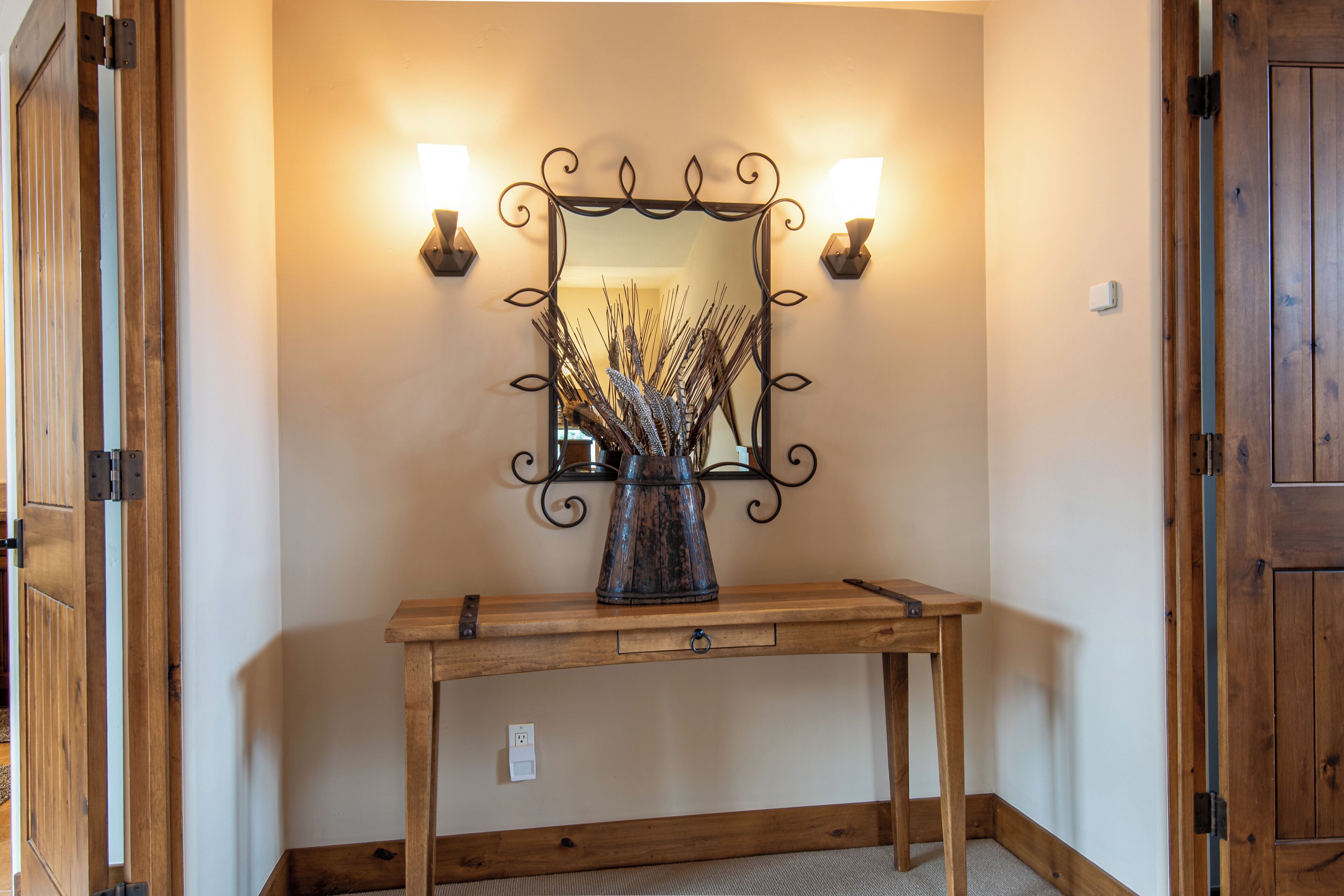 A wooden console table with a mirror above it, flanked by wall sconces, and a vase with dried plants on the table.