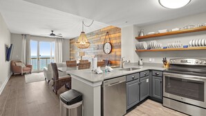 A modern kitchen with a dishwasher, oven, and stainless steel appliances. There is a dining area with wicker furniture and a view of the ocean.