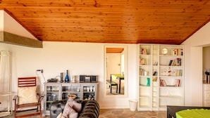 Ein Wohnzimmer mit Holzbalkendecke, Kamin, Bücherregal und Essbereich mit Tisch und Stühlen.