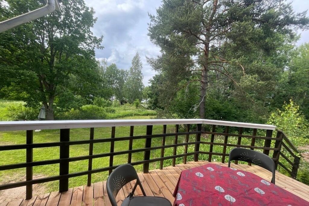 A wooden deck with a table and chairs, surrounded by a black railing, overlooking a grassy area with trees.