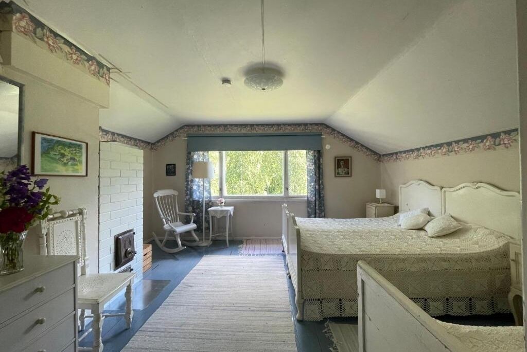 A bedroom with a double bed, a chair, a small table, and a window with a view of greenery.