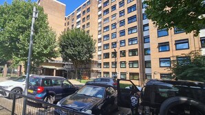 Ein Parkplatz mit mehreren Autos, darunter ein schwarzer SUV und ein weißes Auto, vor einem mehrstöckigen Gebäude mit Backsteinfassaden und zahlreichen Fenstern.