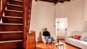 A room with a wooden staircase, a white sofa with a blue and red pillow, a small round table with a remote control, and a white door. - Moinho Pedra Pomes in Santa Cruz da Graciosa (Santa Cruz da Graciosa)