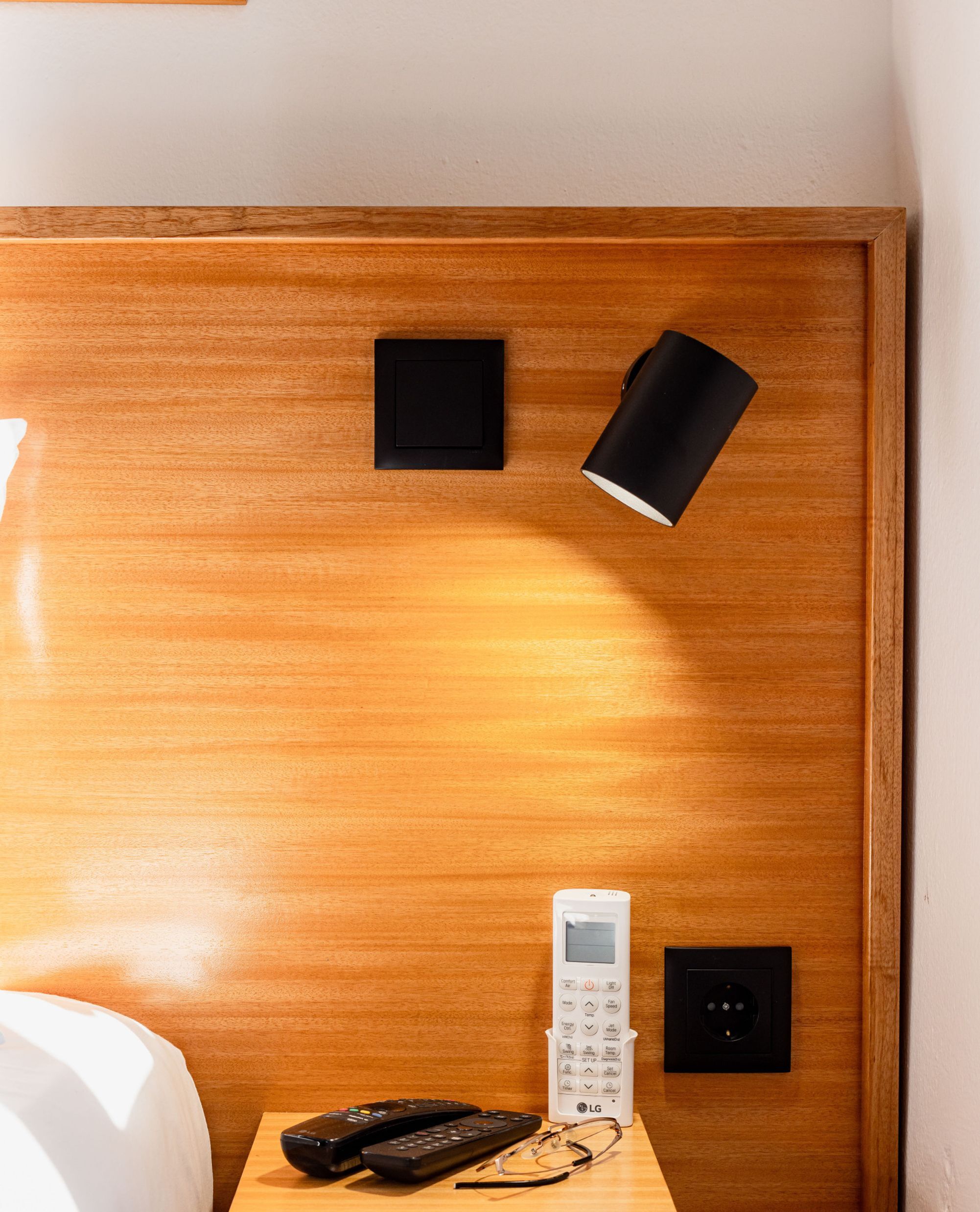 Un mur recouvert de panneaux de bois sur lequel on aperçoit un objet carré noir, un objet cylindrique noir, une télécommande blanche, une télécommande noire et une paire de lunettes posées sur une surface en bois.