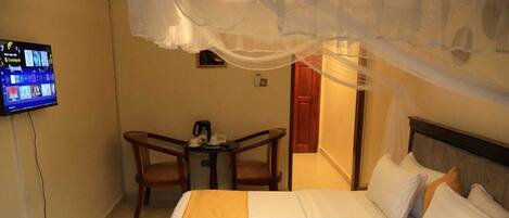 A hotel room with a bed, a TV mounted on the wall, a small table with chairs, and a door leading to another room.
