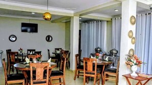 A dining area with wooden tables and chairs, a television mounted on the wall, and a kitchen area visible through a glass partition. - Hotel Casabella (Entebbe)
