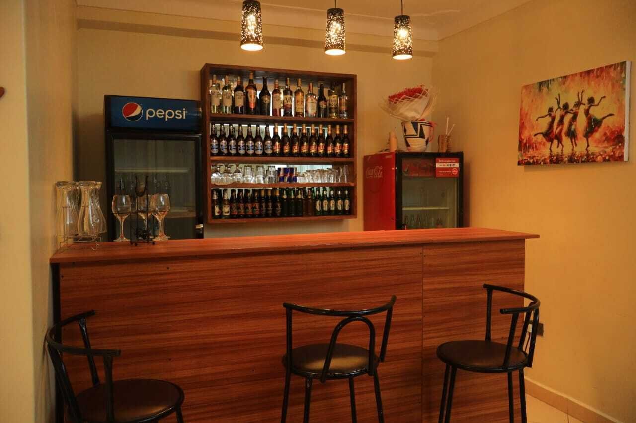 Uma área de bar com um refrigerador da Pepsi, prateleiras com garrafas e um cooler da Coca-Cola. Há três cadeiras e um quadro na parede.