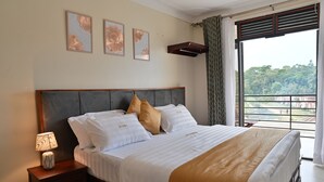 A neatly made bed with white and gold bedding, a bedside lamp, and a nightstand. There are three framed floral artworks on the wall above the bed. - Hotel Casabella (Entebbe)
