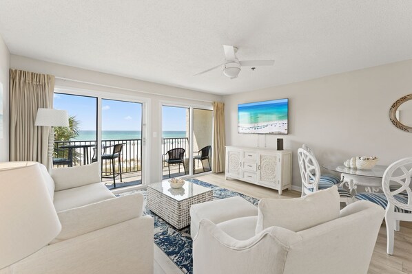A spacious living room with a balcony overlooking the ocean, a flat-screen TV mounted on the wall, and a white dining area with striped cushions.