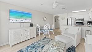 A modern living room with a large TV, white furniture, a dining area with chairs, and a kitchen with appliances. - Windancer 209. Gulf Front Condo, Free Beach Service, Pool, Grill, Sugar Sand! (Miramar Beach)