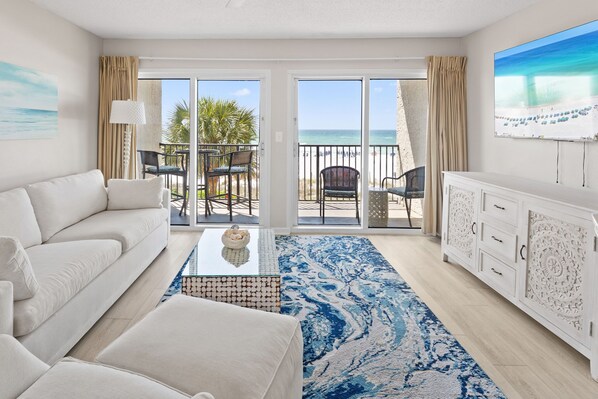 A modern hotel room with a white sofa, a coffee table, a patterned rug, a white cabinet, and a balcony with a view of the beach.