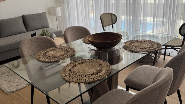 Una sala moderna con una mesa de comedor de vidrio, individuales tejidos, sillas tapizadas en gris, un sofá gris y vistas a una piscina a través de cortinas translúcidas.