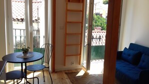 Ein Zimmer mit einem blauen Sofa, einem runden Tisch mit zwei Stühlen, einer kleinen Topfpflanze und einer an der Wand angelehnten Holzleiter.
