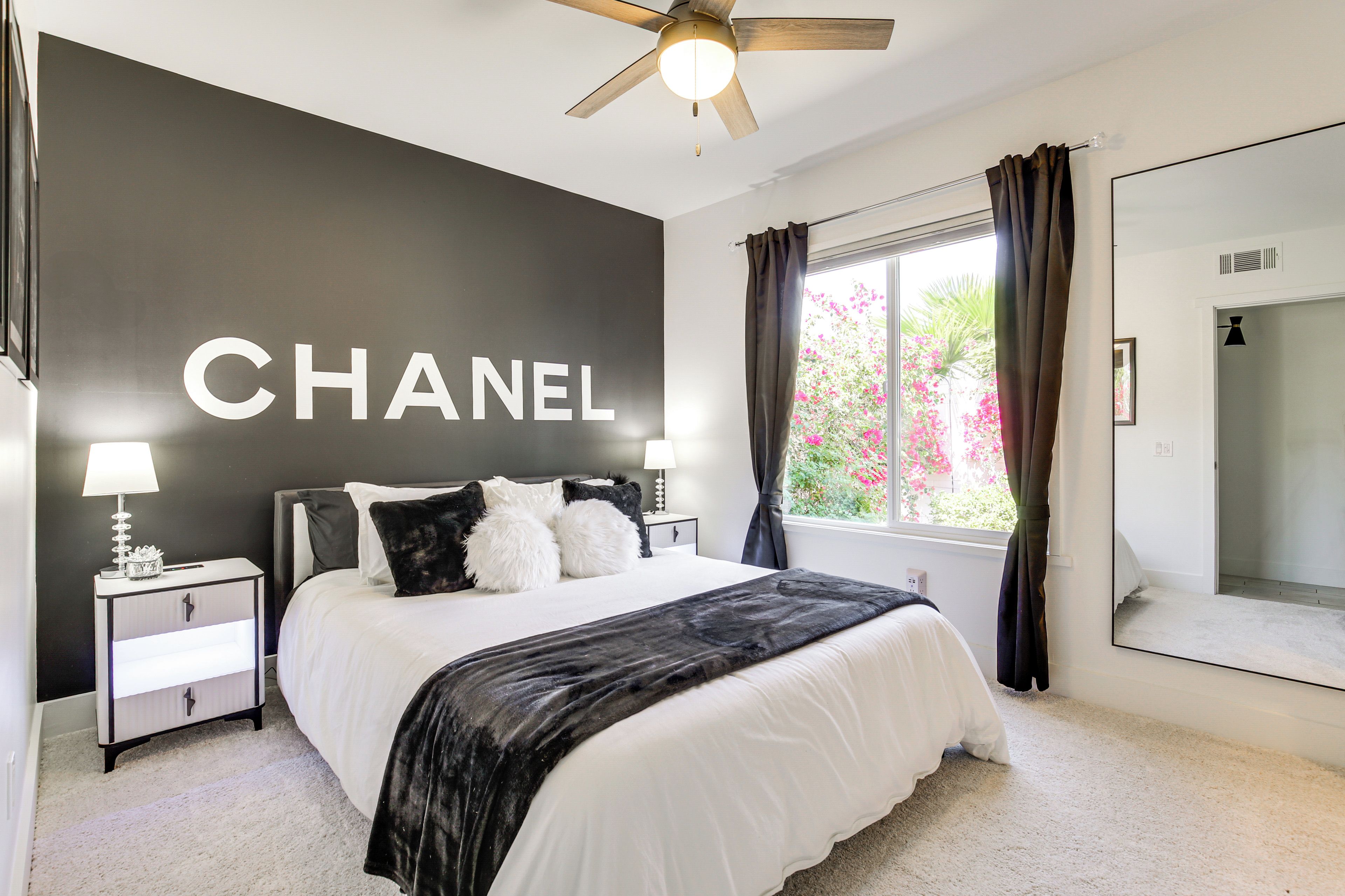 Un dormitorio con una cama grande, una mesita de noche con lámpara, un ventilador de techo, una ventana con cortinas y una pared con la palabra "CHANEL".