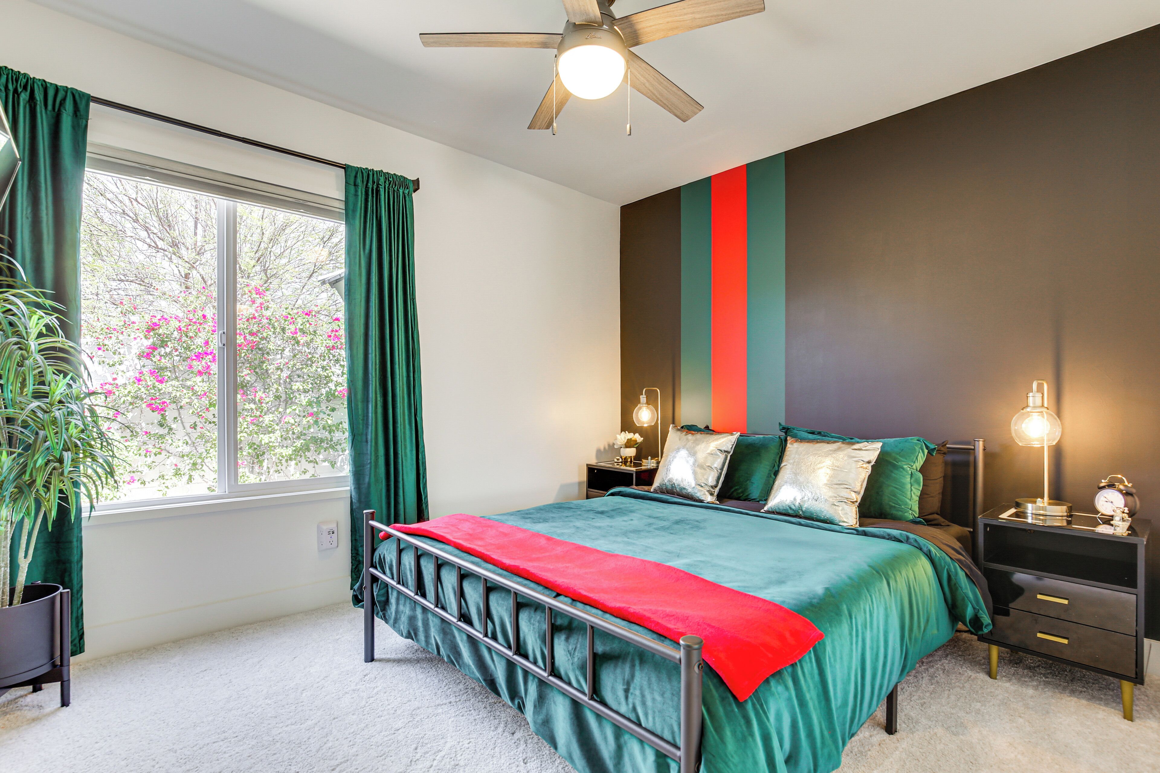 A bedroom with a bed, green curtains, a ceiling fan, a nightstand with a lamp, and a window with a view of pink flowers.
