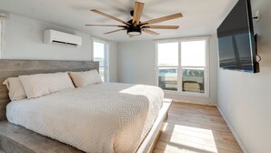 Une chambre à coucher comprenant un lit, un ventilateur de plafond, un climatiseur, une grande fenêtre et un téléviseur fixé au mur.