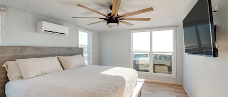A bedroom with a bed, a ceiling fan, an air conditioner, a large window, and a TV mounted on the wall.
