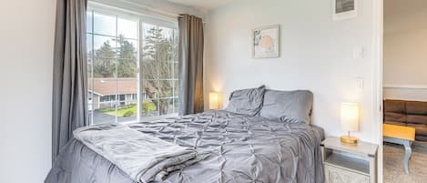 A bedroom with a large bed, a bedside table with a lamp, a window with curtains, and a view of the outside.