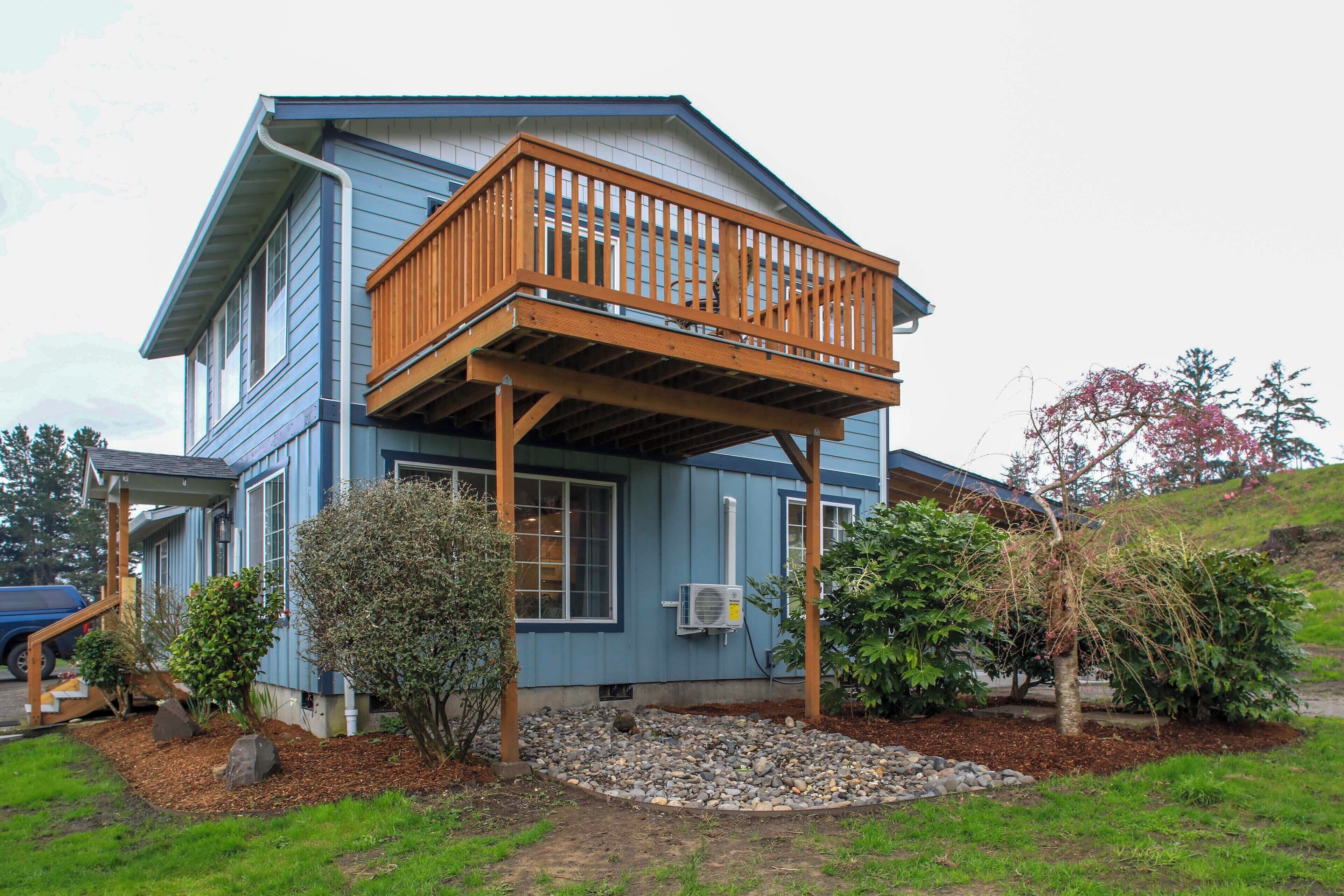 Une maison à deux étages dotée d’un balcon en bois, d’un petit jardin avec des rochers et des arbustes, et d’une façade bleue.