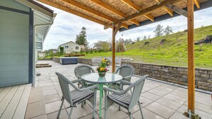 A covered patio with a round table and four chairs, a flower vase, and a view of a house and grassy hill in the background. - 3 Mi to Beach: Oregon North Coast Retreat (Warrenton)