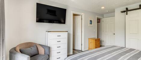 Un dormitorio con una cama, un televisor montado en la pared, un aparador y una puerta que da a otra habitación.