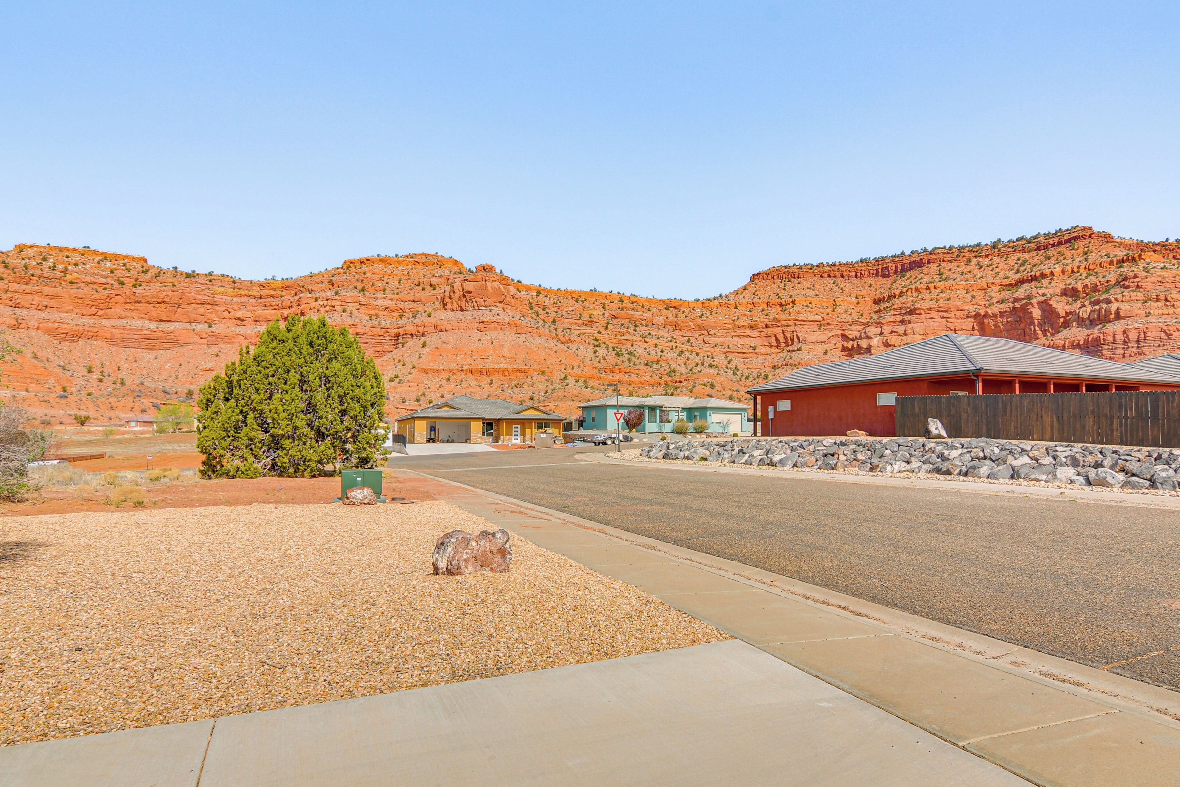 A residential street with houses, a clear sky, and red rock formations in the background.