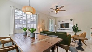 A dining area with a wooden table, chairs, and potted plants. There is a ceiling fan and a TV mounted on the wall.
