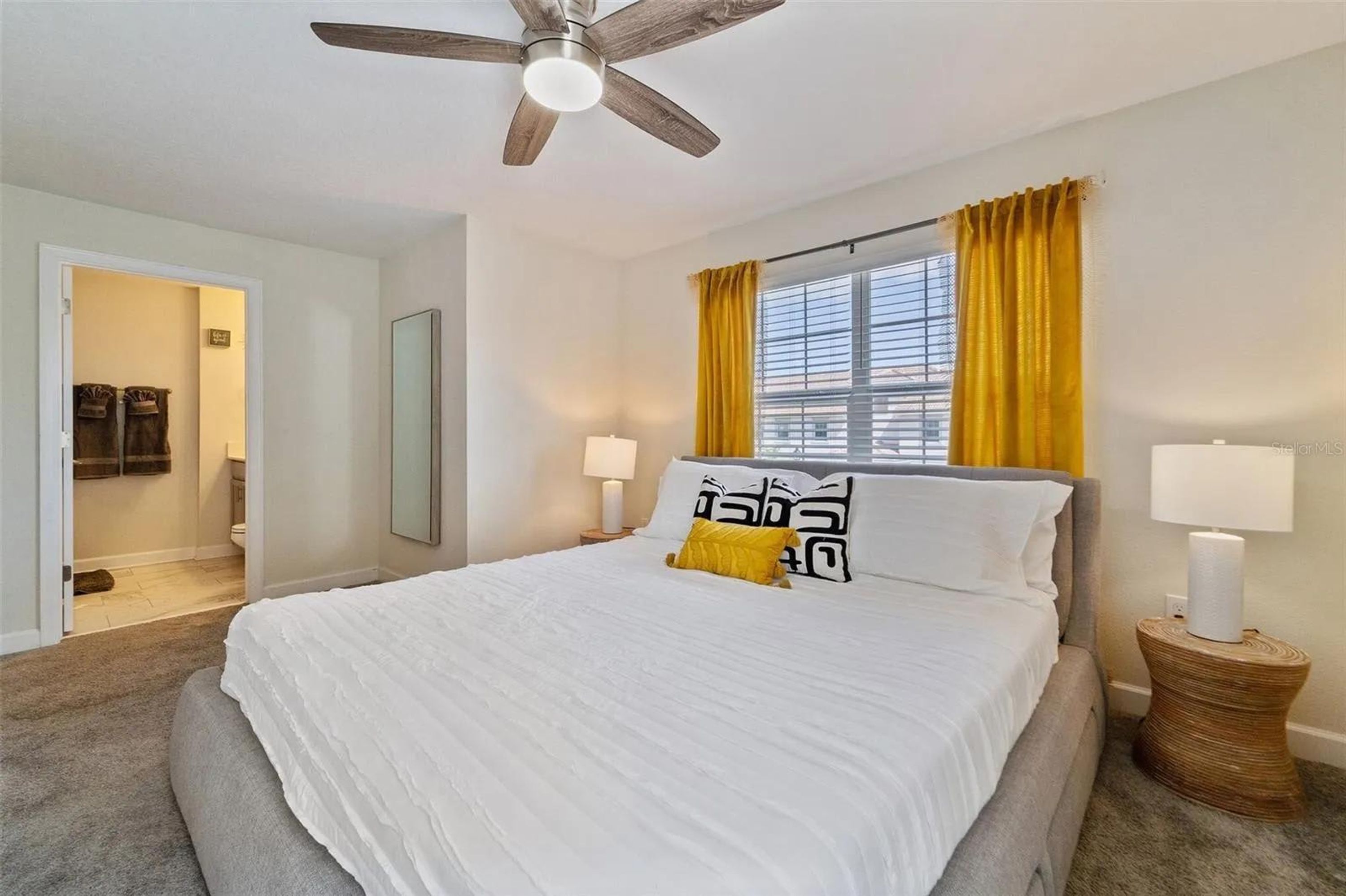 A bedroom with a bed, a ceiling fan, a window with yellow curtains, and a bedside lamp.
