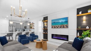 Ein modernes Wohnzimmer mit Kamin, großem Fernseher und einer Küche im Hintergrund.