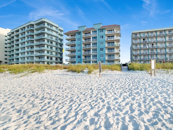 A beachfront hotel with multiple floors, balconies, and a sandy beach in the foreground. - Emerald Key #602 | 3BD/2BA, Pool, Beach Access, Across from Shopping Center (Orange Beach)
