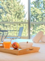 A breakfast tray with a glass of orange juice, an apple, and a bowl on a balcony with a view of trees.