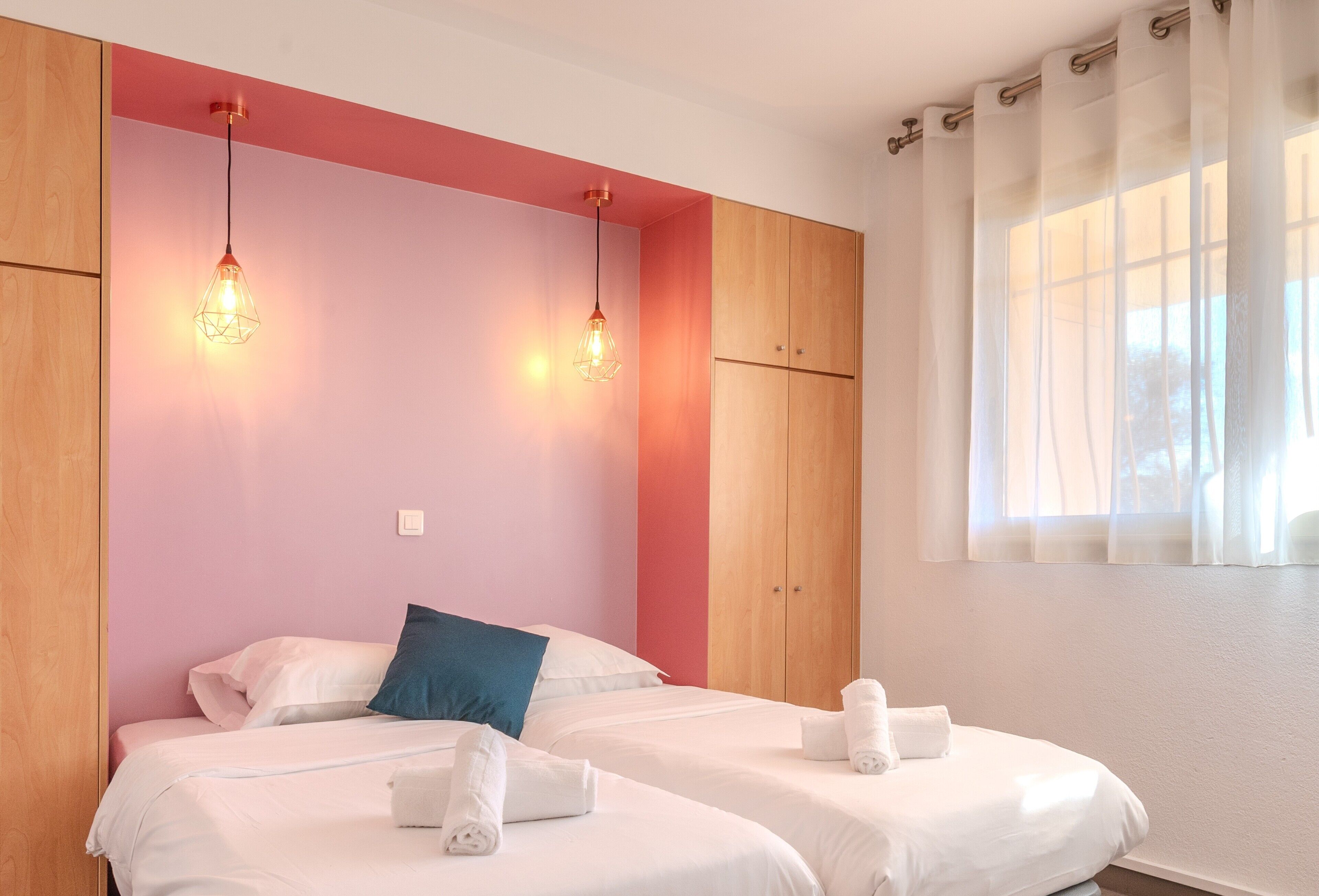 A hotel room with two beds, wooden wardrobe, and a window with sheer curtains.