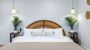 Una cama bien tendida con cabecera de madera, dos mesitas de noche con plantas en macetas y lámparas colgantes.