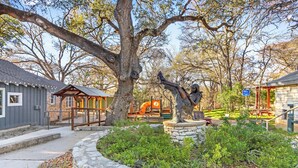 Un espacio al aire libre rústico con un gran árbol, un camino de piedra y un parque infantil con tobogán y columpios.