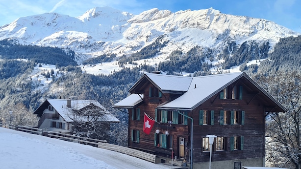 Un bâtiment de style chalet, orné d’un drapeau suisse, entouré de montagnes enneigées et surplombé d’un ciel dégagé.