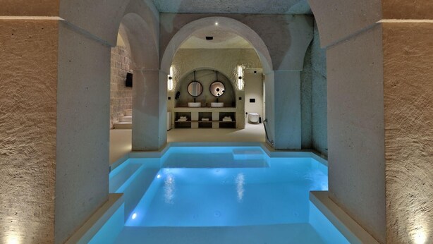 An indoor swimming pool with a stone wall and arched ceiling, leading to a bathroom area with a sink and mirror. - Simurg Cave House (Urgup)