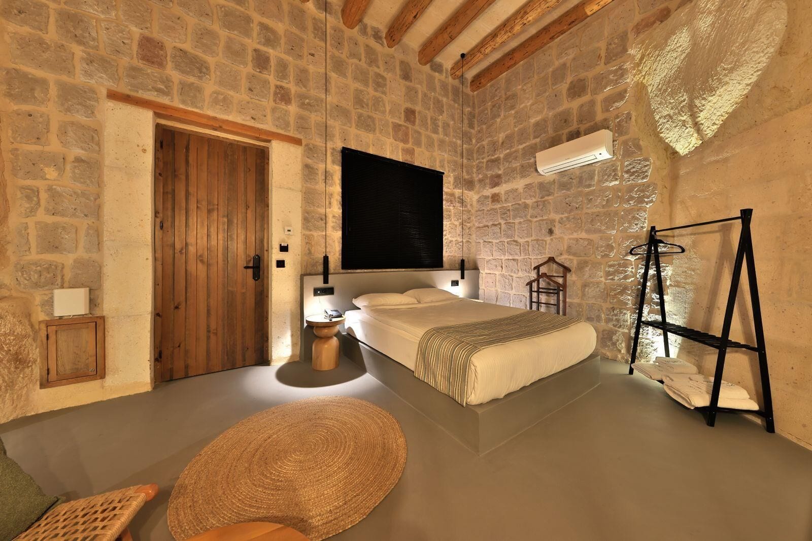 A bedroom with a stone wall, a wooden door, a bed, a bedside table, a hanging light, a black air conditioner, and a black metal rack with clothes.
