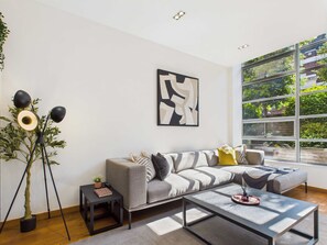 A modern living room with a grey sofa, a coffee table, and a floor lamp. There is a large window with a view of greenery outside. - Refined and Modern Camden Town Apartment (Greater London)