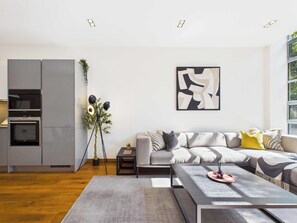A modern living room with a grey sofa, a coffee table, and a floor lamp. There is a kitchen area with built-in appliances and a potted plant.