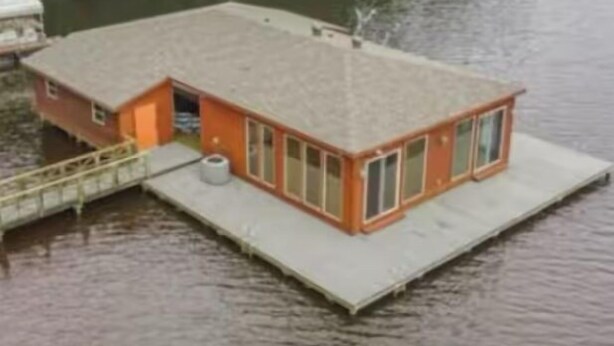 Un petit bâtiment d’un seul étage, doté d’un toit plat, situé sur une plateforme flottante au milieu d’une étendue d’eau, et entouré de verdure.