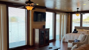 Un salon spacieux avec un grand canapé, une petite table et un téléviseur. De grandes fenêtres sont équipées de stores, et on y trouve également un ventilateur de plafond et un meuble en bois.